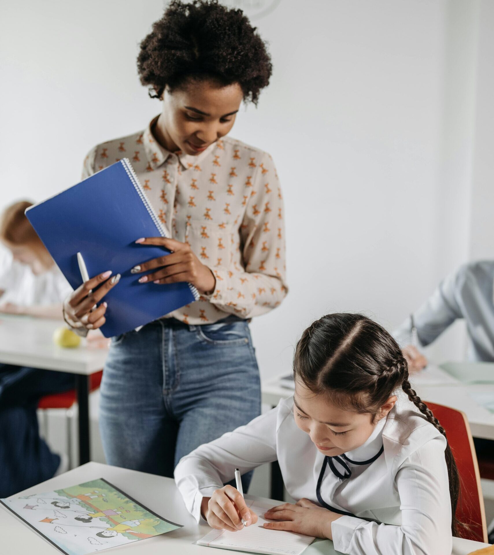 A diverse teacher helps a student with schoolwork in a classroom environment.